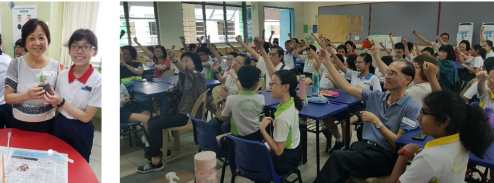Two women smiling while holding a small plant, beside a crowded classroom of students and adults raising their hands.